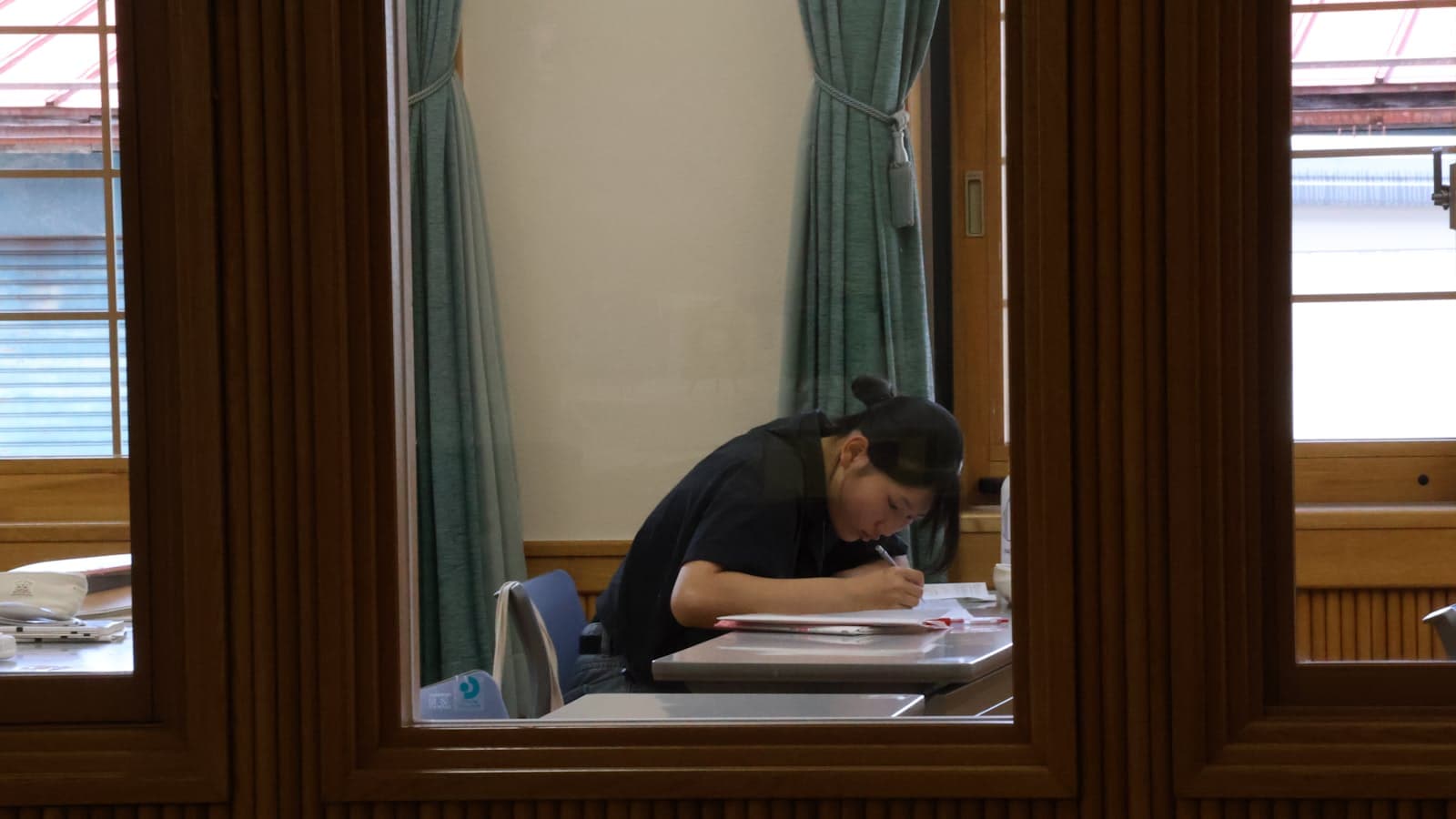 A student writing at a desk by a window, seen through a wooden-framed glass partition