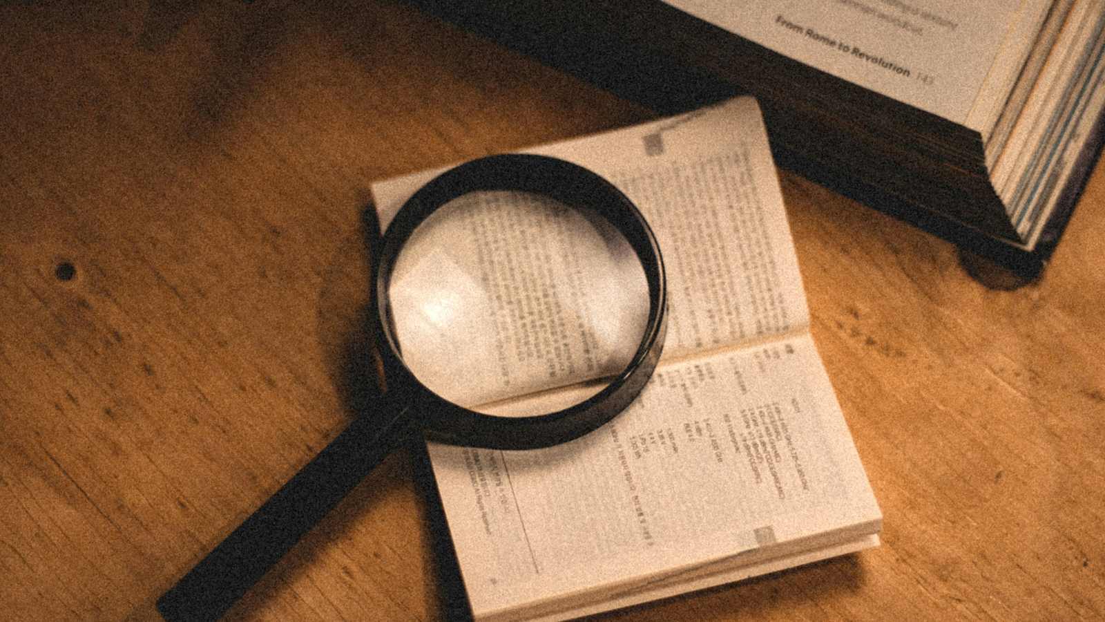 A magnifying glass resting on an open book of small print, beside a larger open book on a wooden table