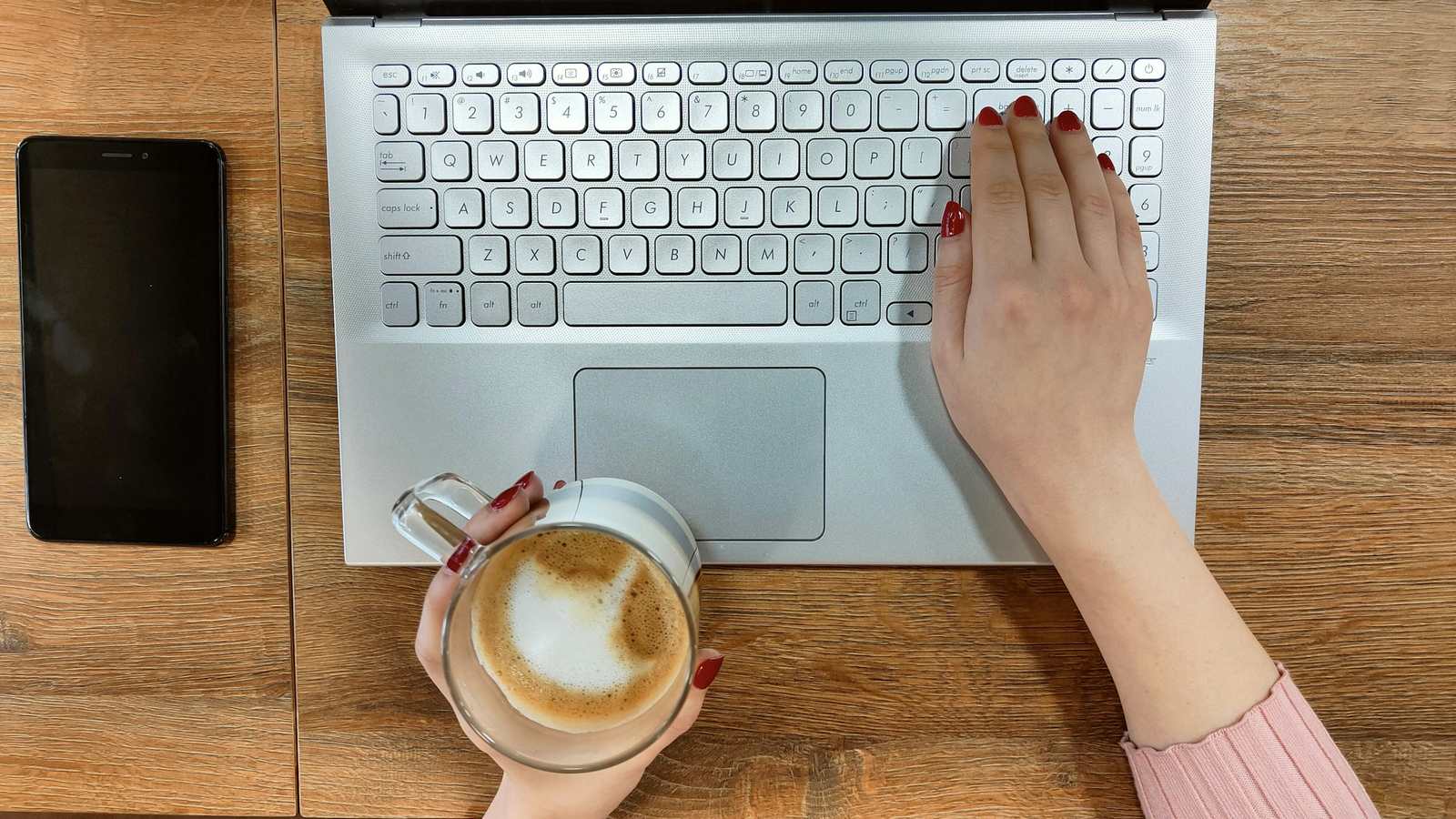 Overhead view of hands typing on a silver laptop and holding a glass of latte, beside a smartphone on a wooden desk